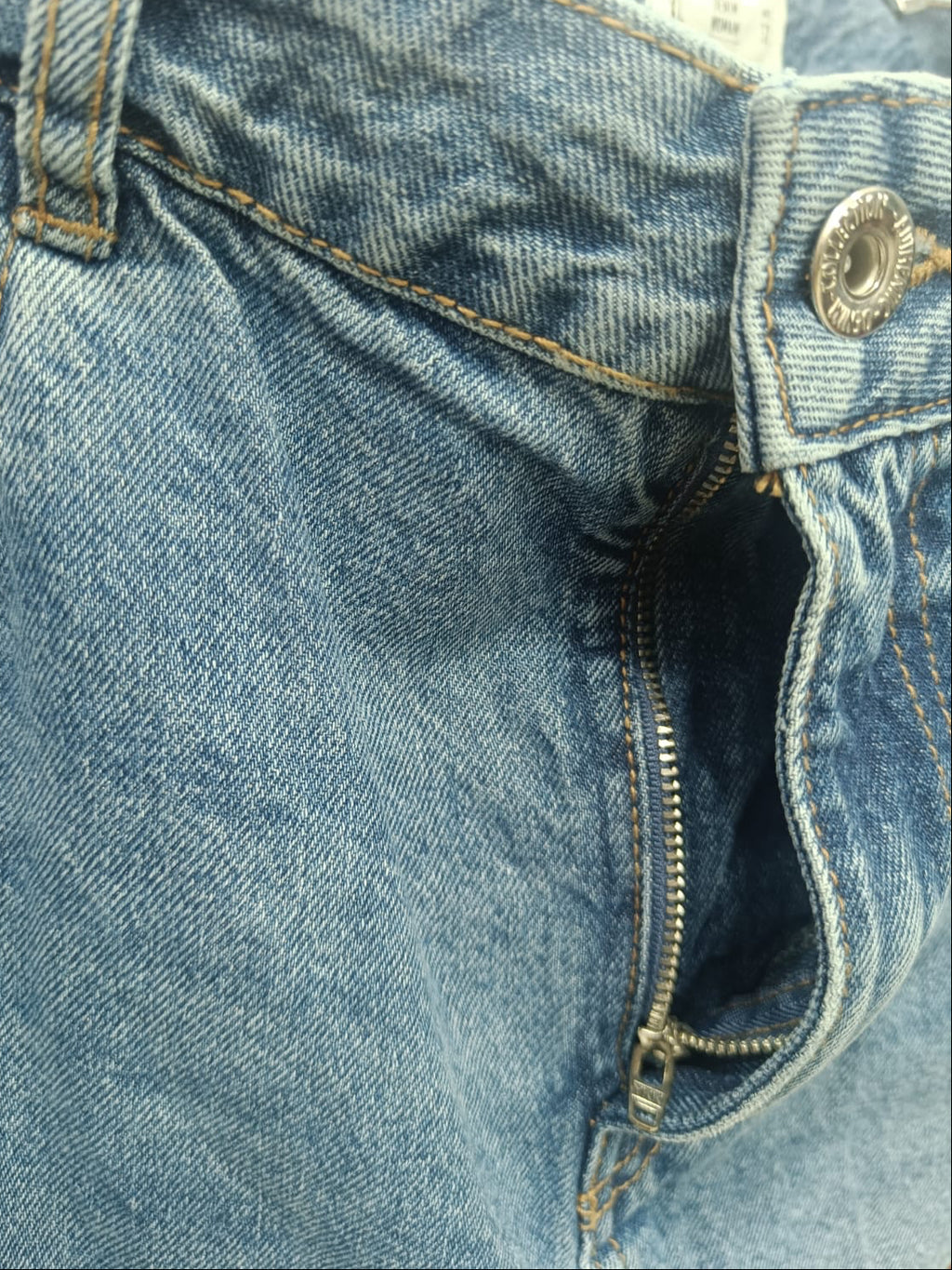 Close-up detail of a pair of blue denim jeans showing the waistband, button closure, zipper pocket, and stitching.