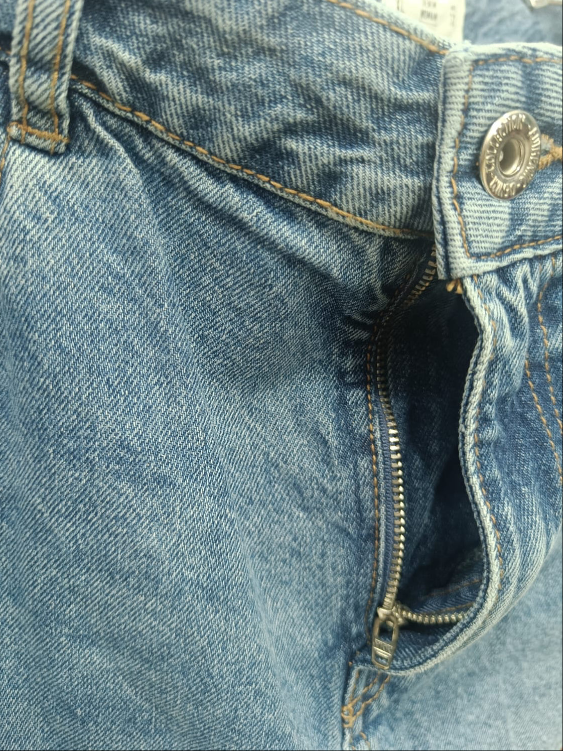 Close-up detail of a pair of blue denim jeans showing the waistband, button closure, zipper pocket, and stitching.