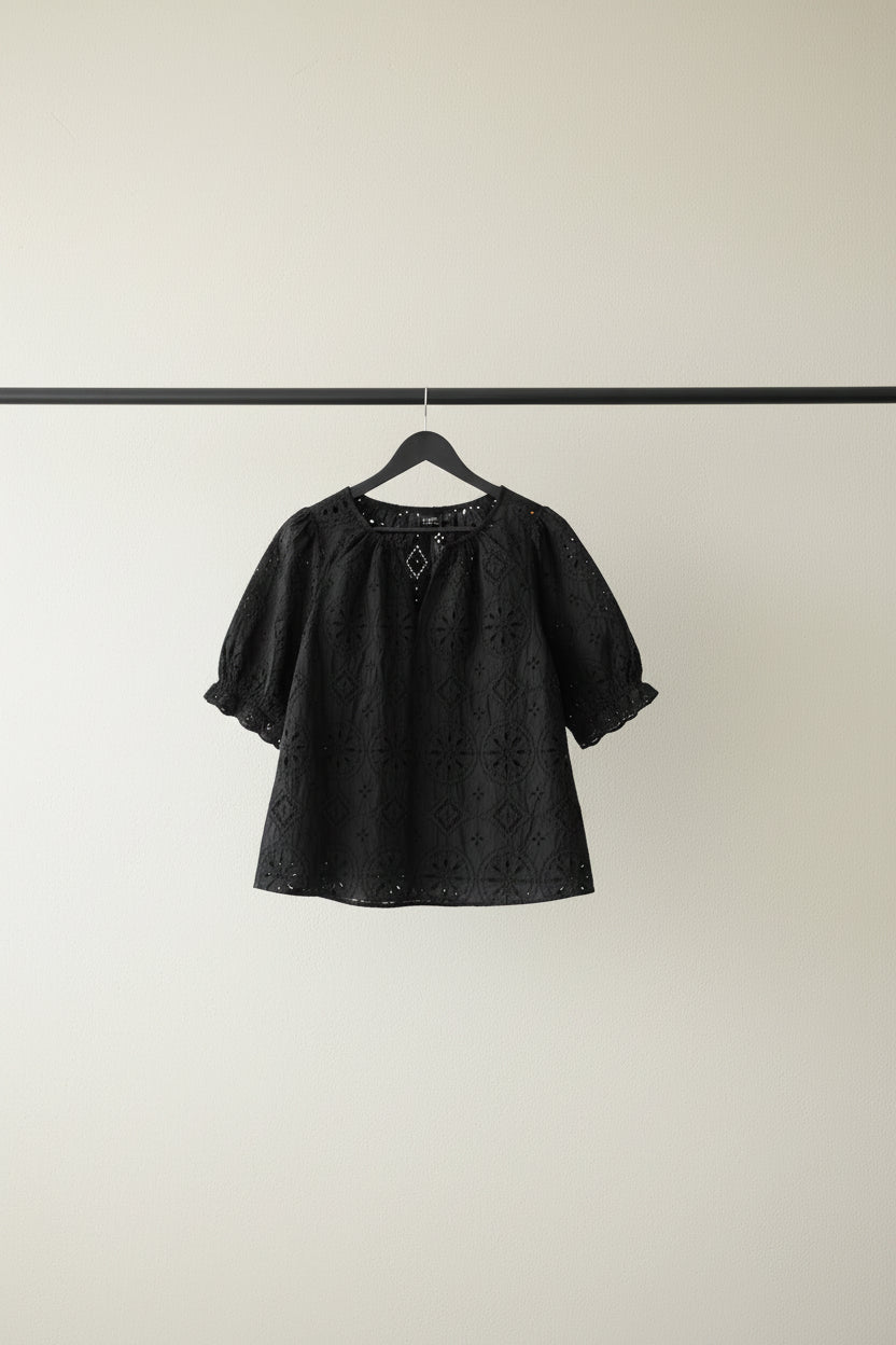 A black short-sleeve eyelet lace top hanging on a wooden hanger from a tree branch outdoors, with green leaves and bright sunlight in the background.