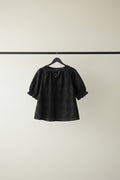 A black short-sleeve eyelet lace top hanging on a wooden hanger from a tree branch outdoors, with green leaves and bright sunlight in the background.