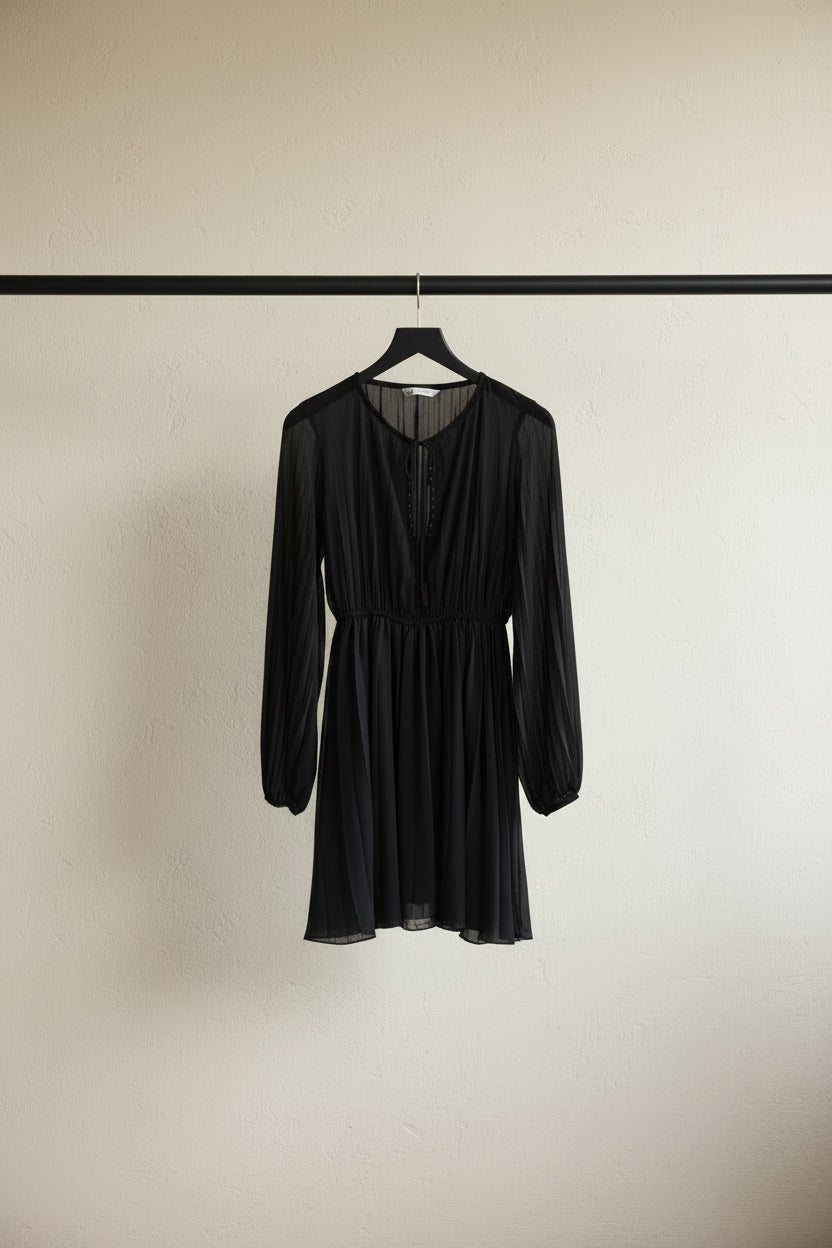 A black long-sleeve pleated dress with a tie-neck detail and gathered waist, hanging on a wooden hanger against a white fabric backdrop.