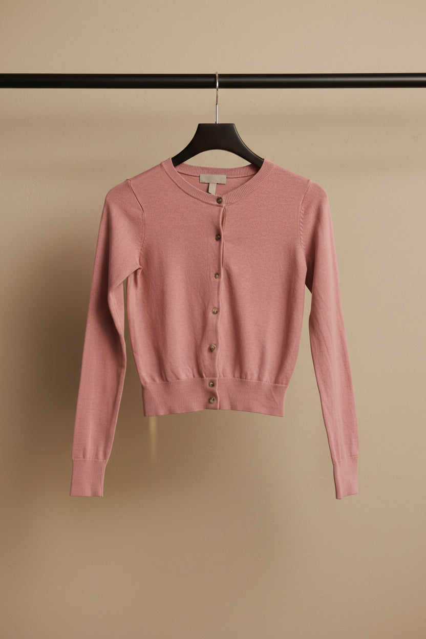 A dusty pink long-sleeve knit cardigan sweater hanging on a wooden hanger against a white fabric backdrop.
