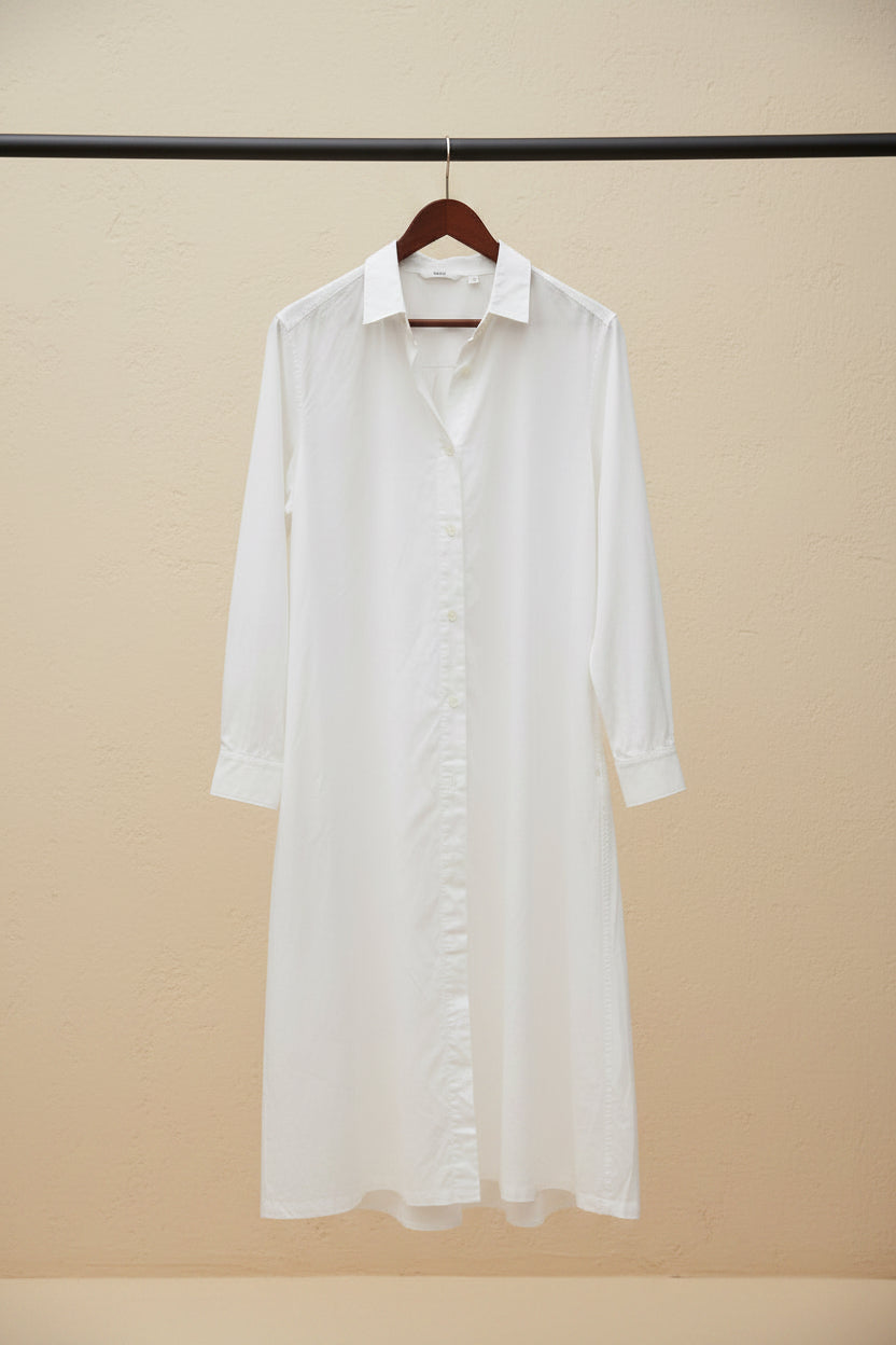 A long white button-front shirt dress with a collar and long sleeves, hanging on a wooden hanger outdoors against a building wall and tree trunk.