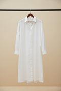 A long white button-front shirt dress with a collar and long sleeves, hanging on a wooden hanger outdoors against a building wall and tree trunk.