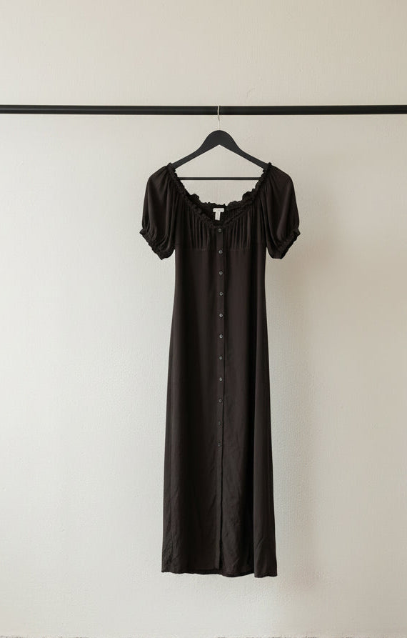 A black midi-length dress with short puff sleeves, a ruffled square neckline, and a button-front closure, hanging on a wooden hanger against a wrinkled white fabric backdrop.