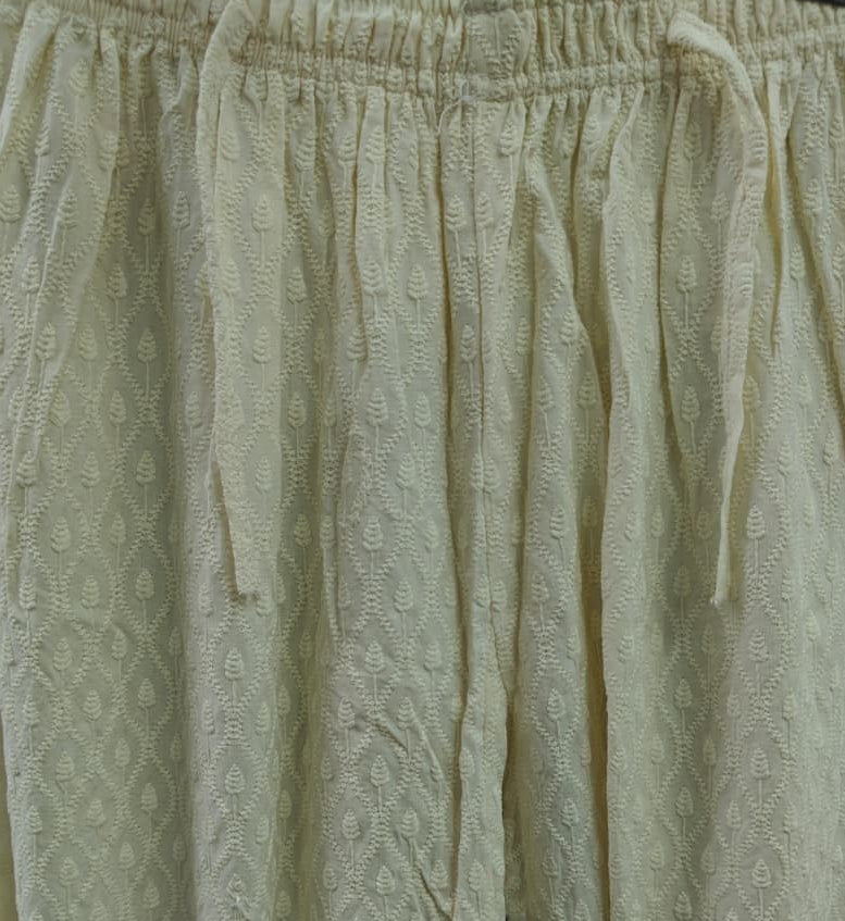 Cream-colored textured knit pants with an elastic drawstring waistband, shown hanging against a dark background.