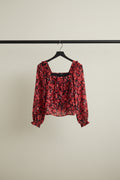 A red and black floral print long-sleeve blouse with a square neckline and ruffled cuffs, laid flat on a white fabric background.