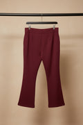 A pair of burgundy cropped pants hanging from a wooden hanger against a wrinkled white fabric backdrop.