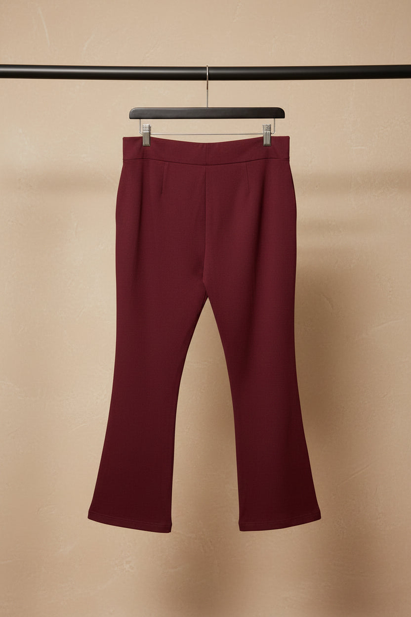 A pair of burgundy cropped pants hanging from a wooden hanger against a wrinkled white fabric backdrop.