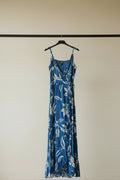 A blue maxi dress with a white floral and leaf pattern, featuring thin spaghetti straps and a ruffled front hem, laid flat on a white fabric background.