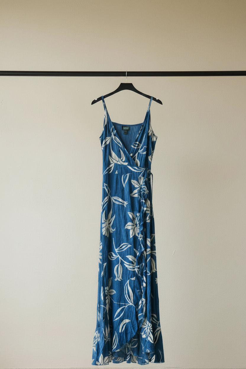 A blue maxi dress with a white floral and leaf pattern, featuring thin spaghetti straps and a ruffled front hem, laid flat on a white fabric background.