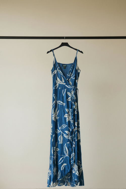 A blue maxi dress with a white floral and leaf pattern, featuring thin spaghetti straps and a ruffled front hem, laid flat on a white fabric background.