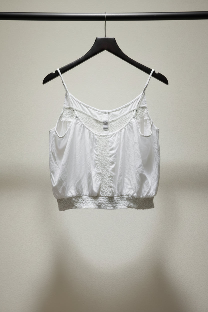 A white lace-trimmed camisole top hanging from a wooden hanger clipped to tree branches outdoors.
