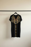A black short-sleeve V-neck dress with a geometric patterned yoke and center panel, hanging on a wooden hanger against a white fabric backdrop.