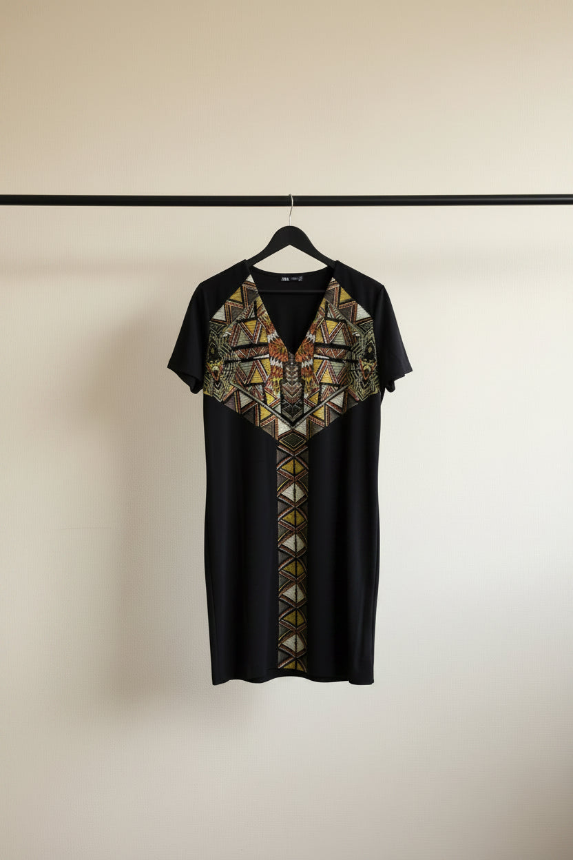 A black short-sleeve V-neck dress with a geometric patterned yoke and center panel, hanging on a wooden hanger against a white fabric backdrop.