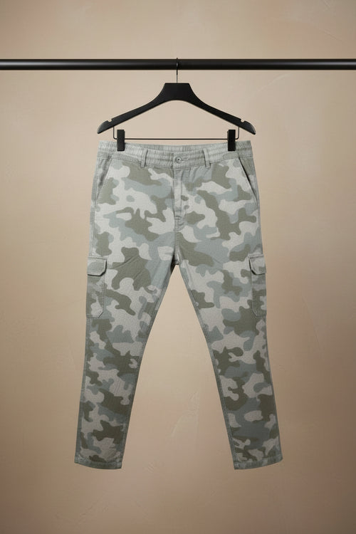 A pair of camouflage cargo pants in shades of beige, gray, and brown, hanging on a dark hanger against a light gray fabric backdrop.