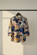 A long-sleeve button-up shirt with a multicolor camouflage pattern in navy blue, mustard yellow, gray, and dusty pink, laid flat on a white fabric background.