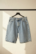 Light wash blue denim shorts hanging from a wooden hanger against a red background.