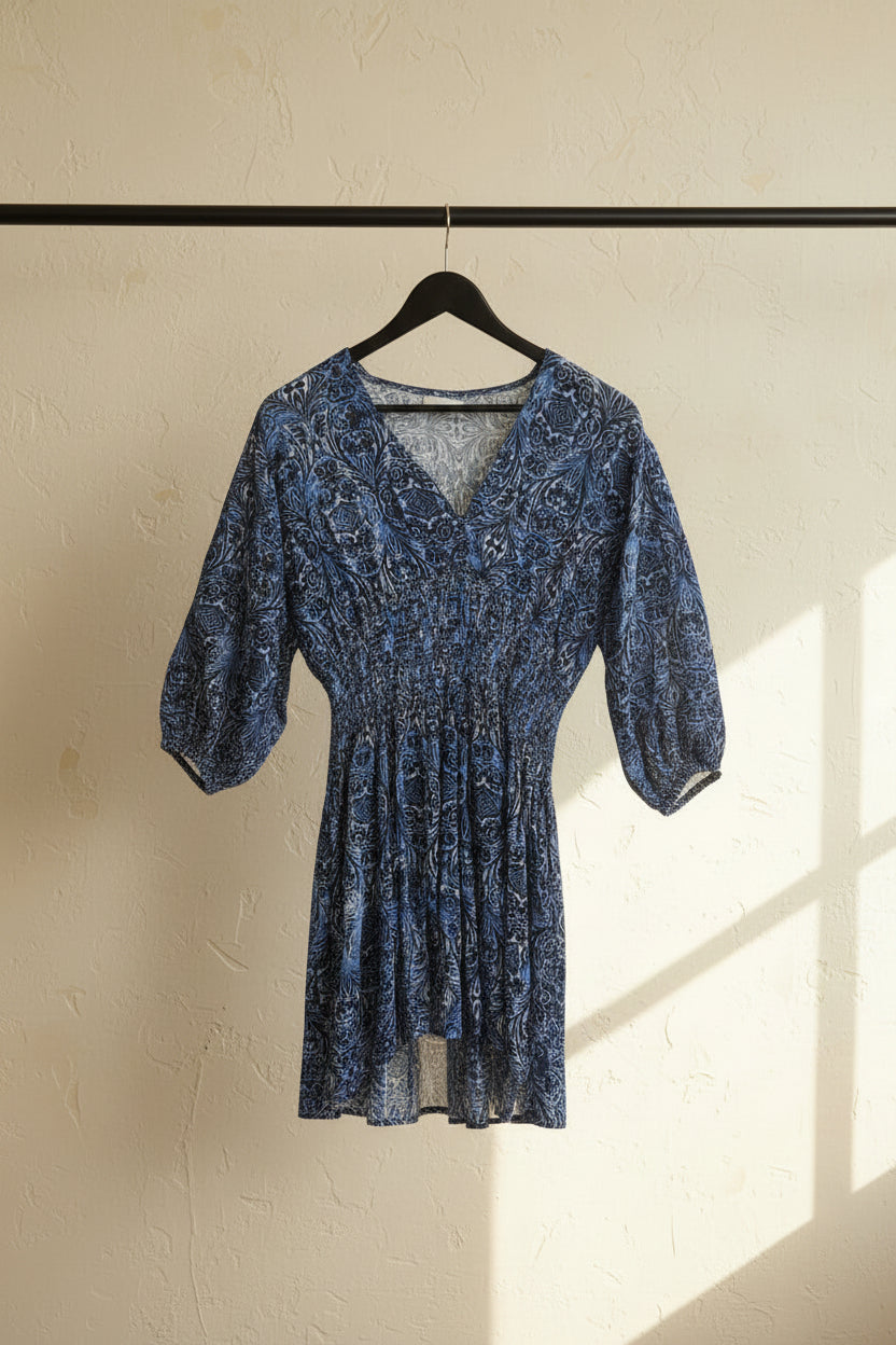 A blue and white patterned dress with three-quarter puff sleeves and a smocked waist, hanging on a wooden hanger against a white fabric backdrop.