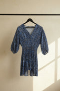 A blue and white patterned dress with three-quarter puff sleeves and a smocked waist, hanging on a wooden hanger against a white fabric backdrop.