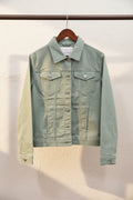 A light sage green denim jacket hanging on a wooden hanger against a textured off-white wall.