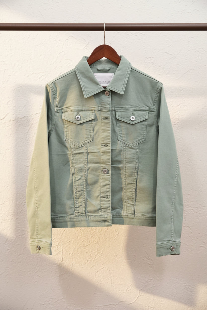 A light sage green denim jacket hanging on a wooden hanger against a textured off-white wall.