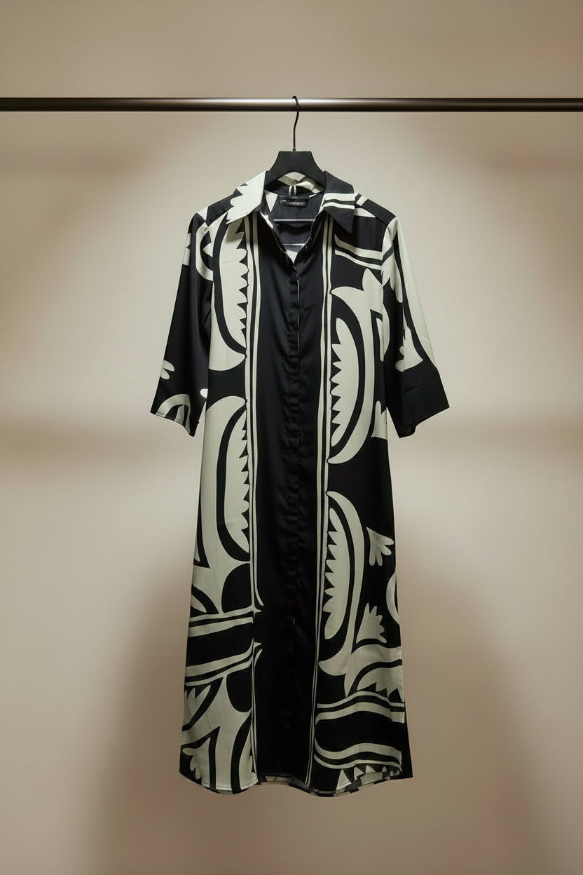 A black and cream abstract patterned shirt dress hanging on a wooden hanger against a white fabric backdrop.