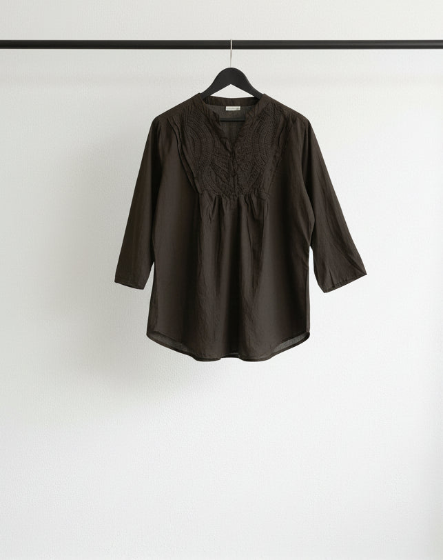A dark brown three-quarter sleeve blouse with a lace-trimmed round neckline, hanging on a wooden hanger against a white fabric backdrop.