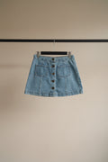 A light blue denim mini skirt with a button-front closure and two front patch pockets, hanging on a wooden hanger against a white fabric background.