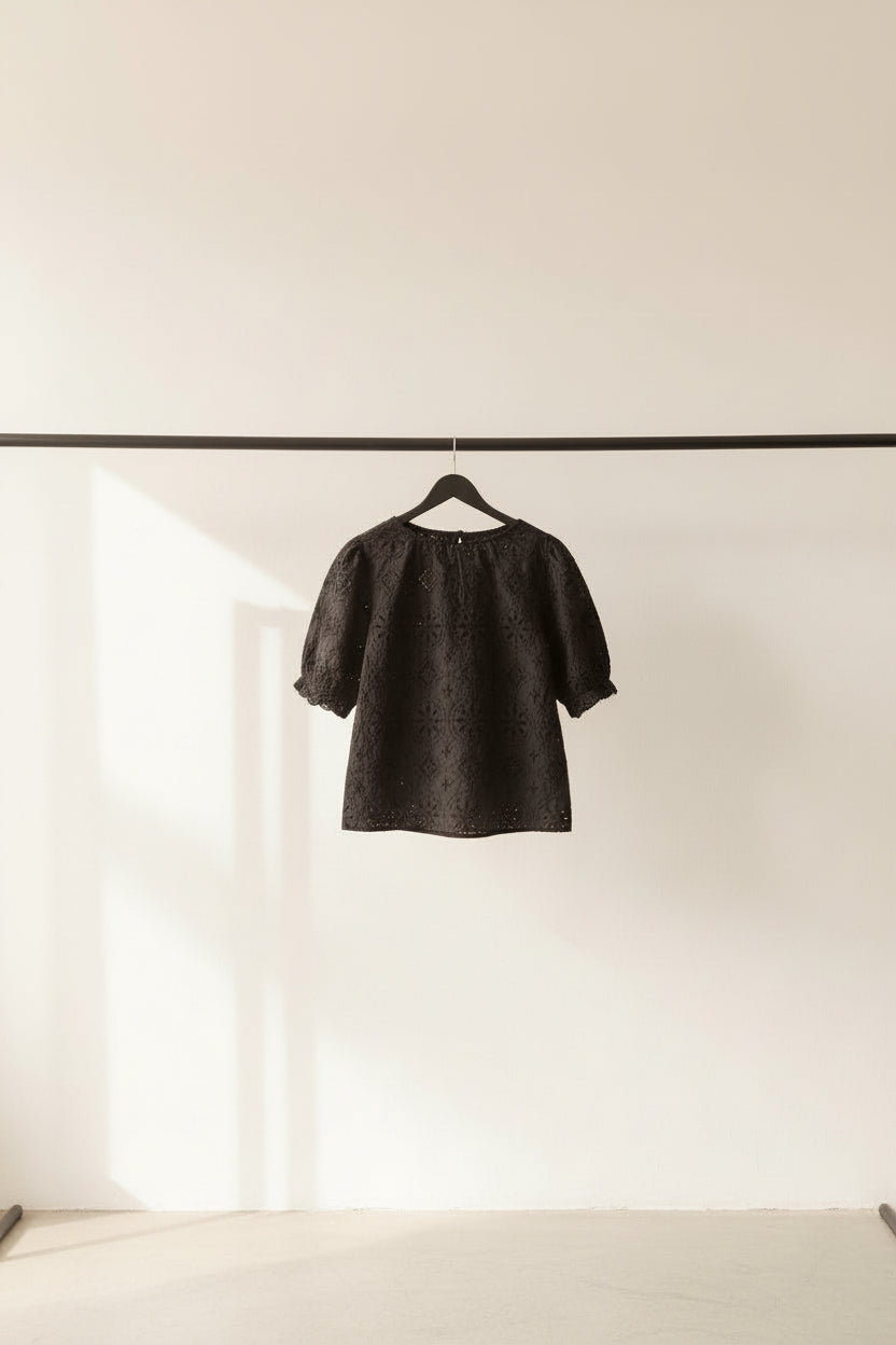 A black embroidered lace blouse with short sleeves and a round neckline, hanging on a wooden hanger outdoors under green leaves.