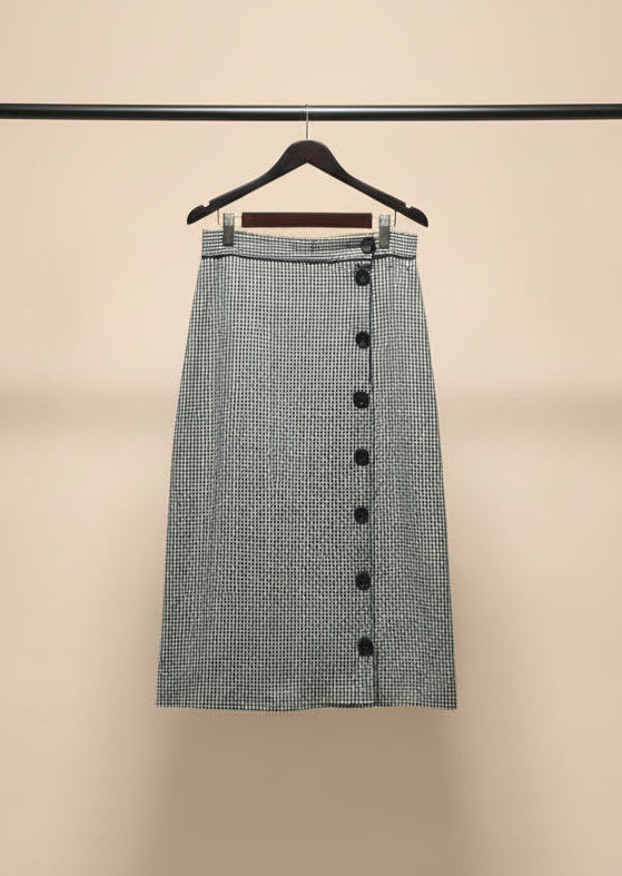 A black and white micro-checkered midi skirt with a button-down front, hanging on a wooden hanger outdoors on a tree branch.