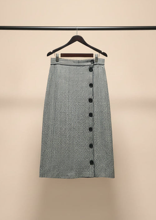 A black and white micro-checkered midi skirt with a button-down front, hanging on a wooden hanger outdoors on a tree branch.