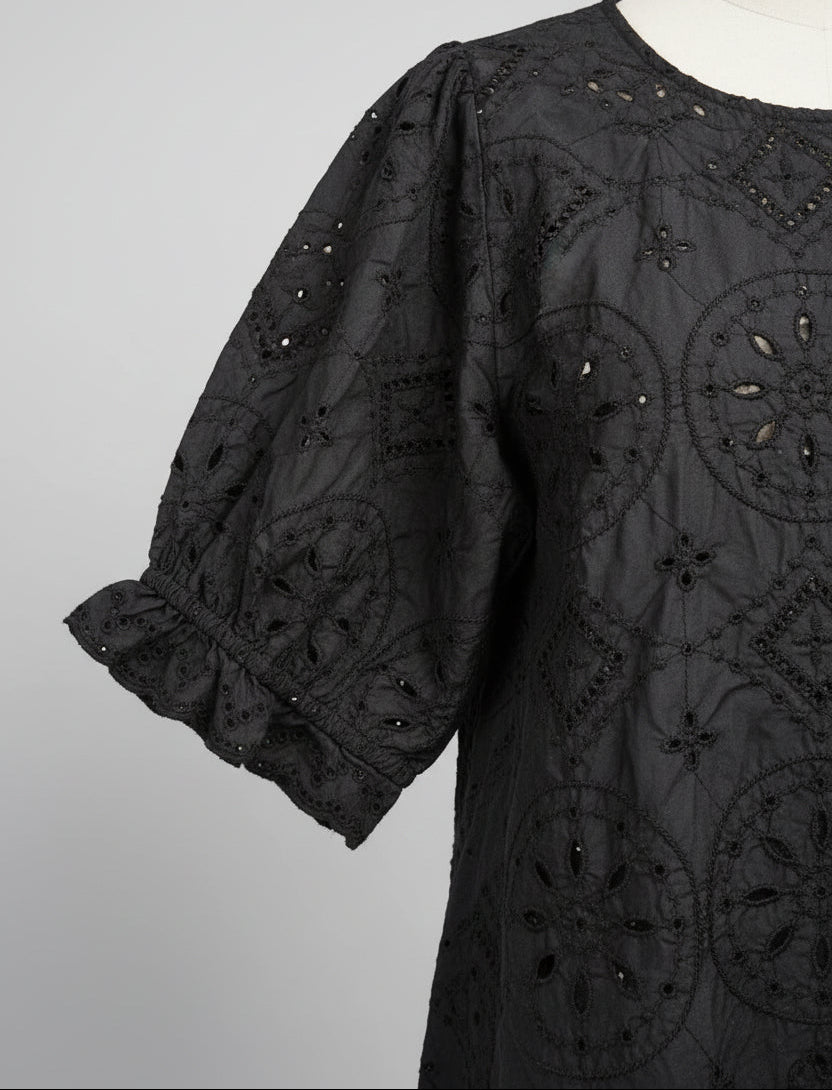 A close-up of a dark brown embroidered lace garment, likely a dress or tunic, featuring intricate floral embroidery and bead embellishments, shown outdoors with green leaves and a wall in the background.