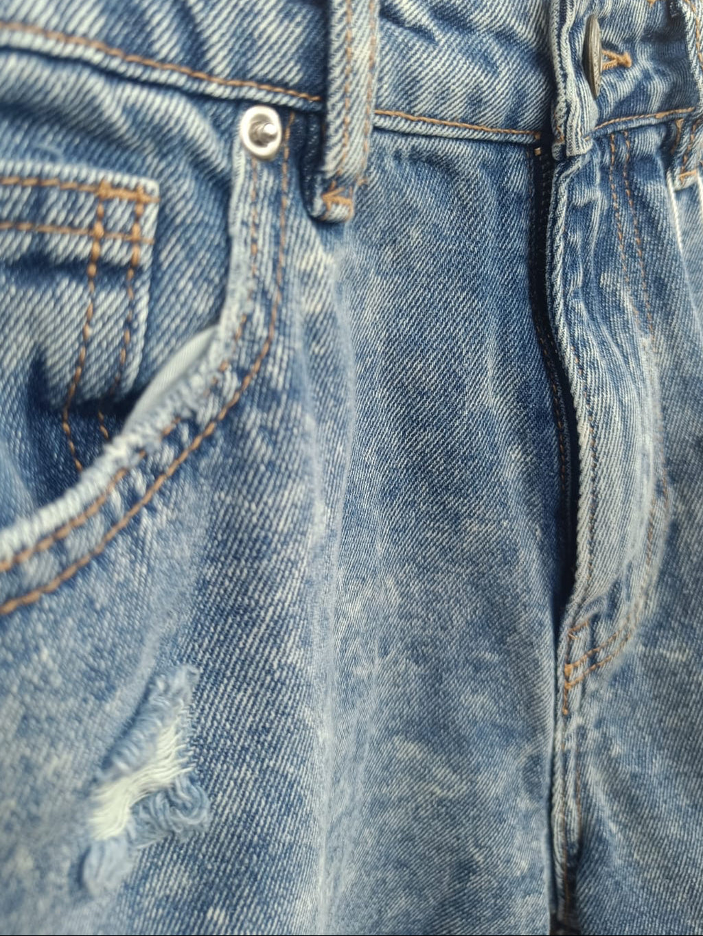 Close-up of a pair of blue denim jeans showing the waistband, belt loops, front pocket, rivet, and a small distressed area near the upper left thigh.