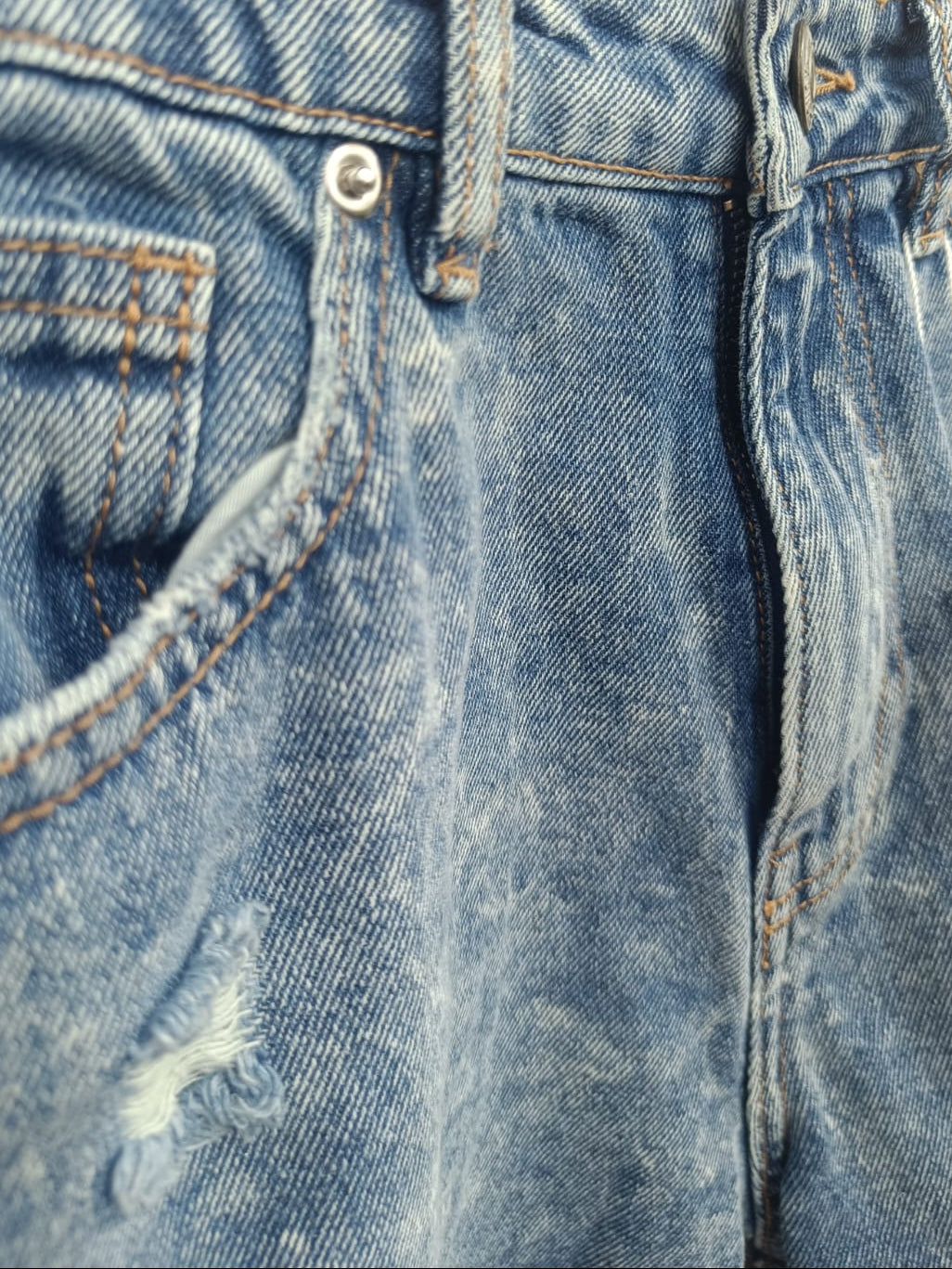 Close-up of a pair of blue denim jeans showing the waistband, belt loops, front pocket, rivet, and a small distressed area near the upper left thigh.
