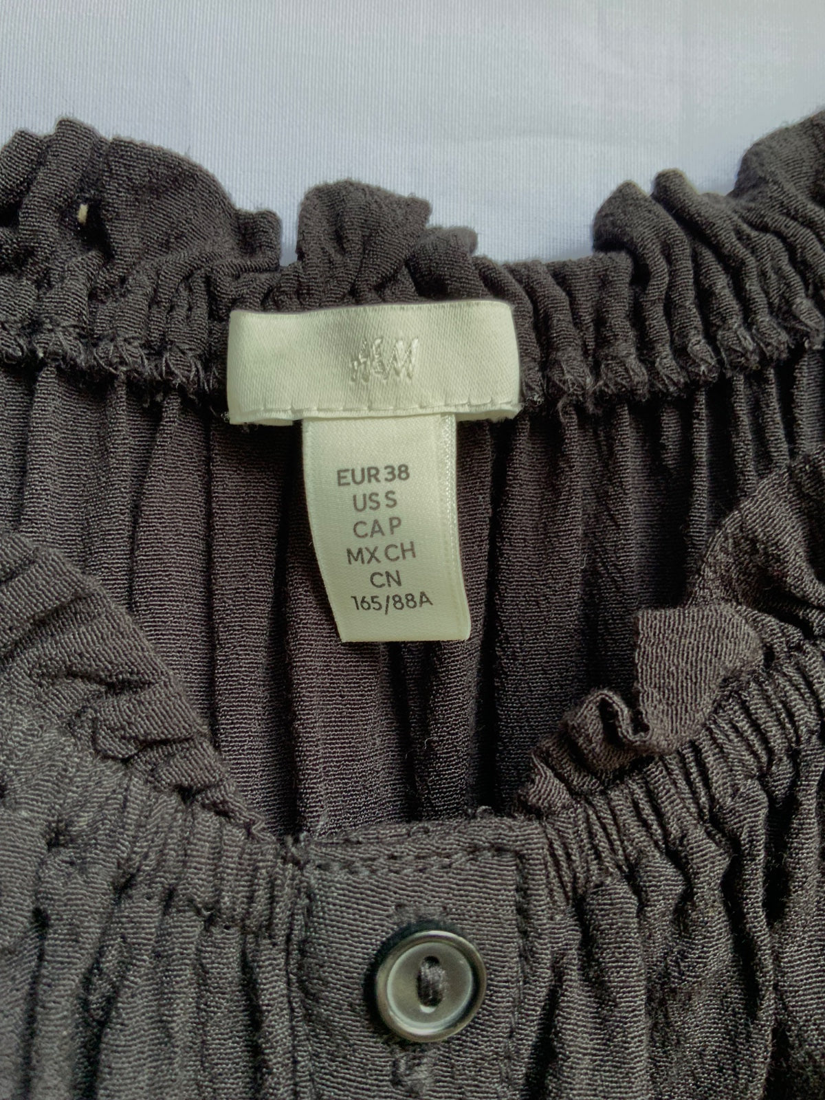 Close-up of a dark brown ruffled top with a cream-colored size tag showing measurements and a metal snap button at the neckline.