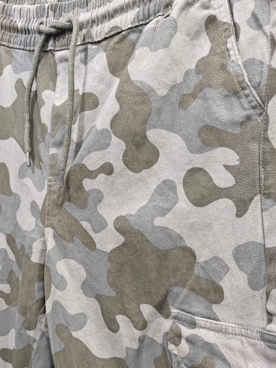 Camouflage cargo pants in a light gray, beige, and brown camo pattern, hanging on a black hanger against a light gray wall.