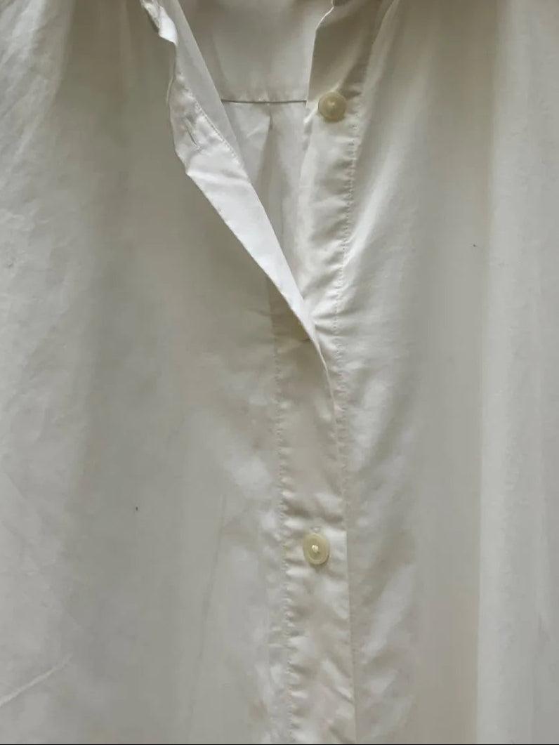 A white long-sleeve button-up shirt hanging on a light wood hanger, with a collar and front buttons visible. The background shows dark tree branches and a light wall.