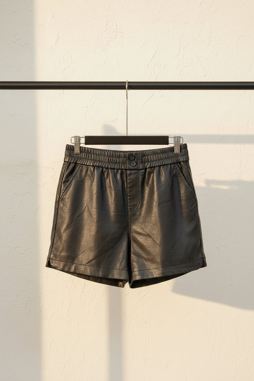 Black faux leather shorts with an elastic waistband and button closure, hanging on a wooden hanger against a red background.