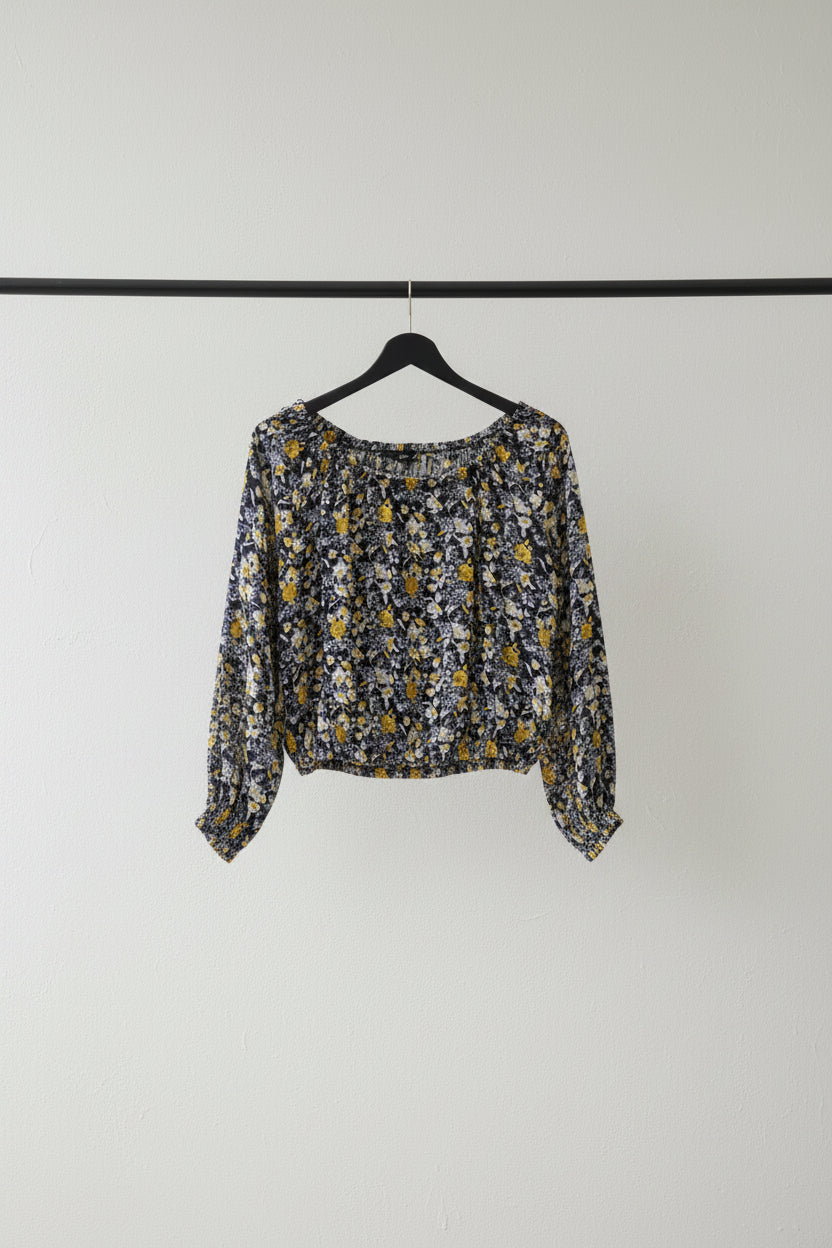 A long-sleeve cropped blouse with a dense yellow, white, and blue floral print on a dark navy background, hanging on a wooden hanger from tree branches.