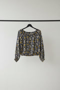 A long-sleeve cropped blouse with a dense yellow, white, and blue floral print on a dark navy background, hanging on a wooden hanger from tree branches.