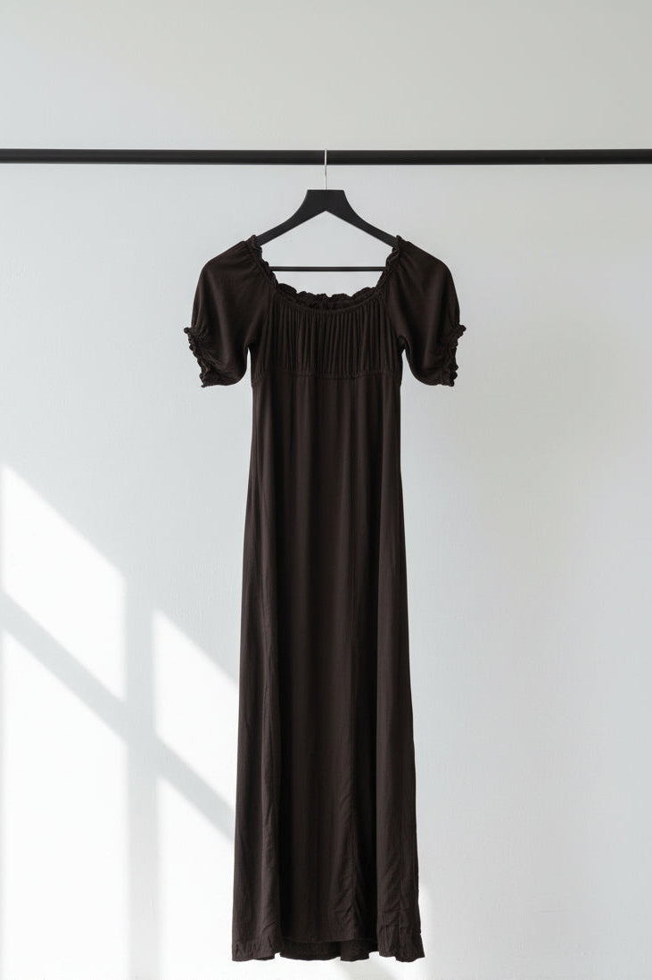 A black maxi dress with short puff sleeves, a square neckline, and a gathered bodice, hanging on a wooden hanger against a white fabric backdrop.