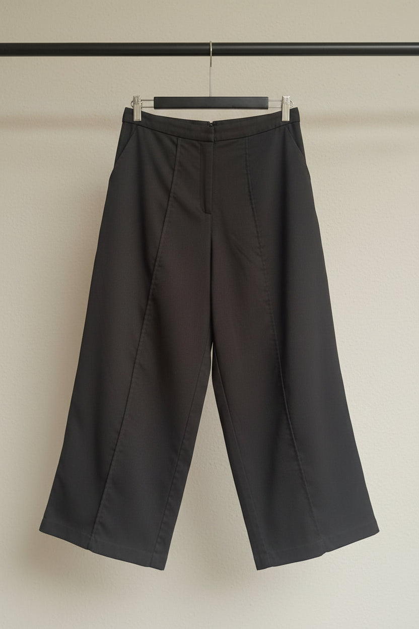 A pair of dark gray cropped wide-leg pants hanging on a wooden hanger against a white fabric backdrop.