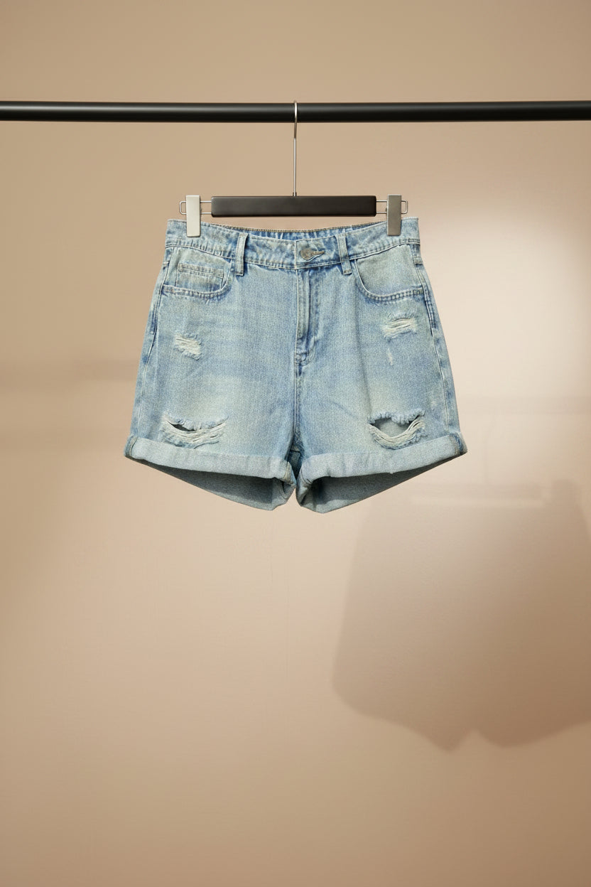 Light wash blue distressed denim shorts with rolled hems, displayed on a wooden hanger against a solid red background.