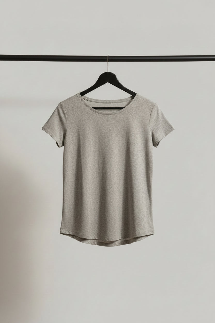 A heather gray short-sleeve t-shirt hanging on a wooden hanger outdoors on a tree branch, with bright sunlight and green leaves creating a backlit effect.