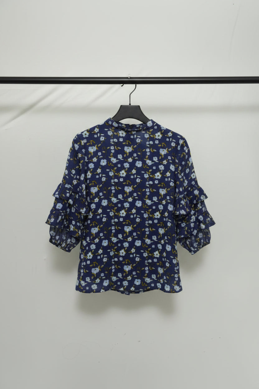 A navy blue long-sleeve blouse with a dense white and light blue floral print and small yellow accents, laid flat on a light-colored fabric background.