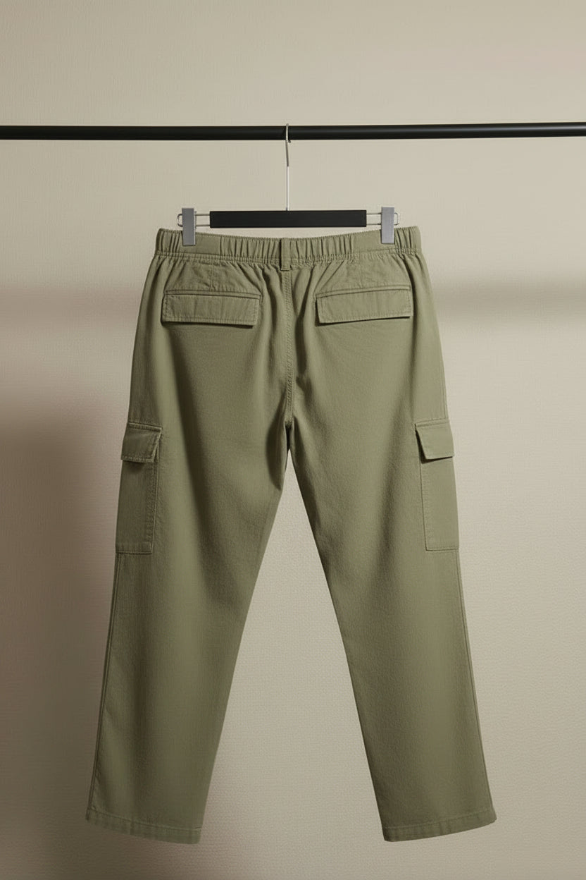 A pair of olive green cargo pants hanging against a gray fabric backdrop, featuring an elastic waistband and multiple utility pockets.