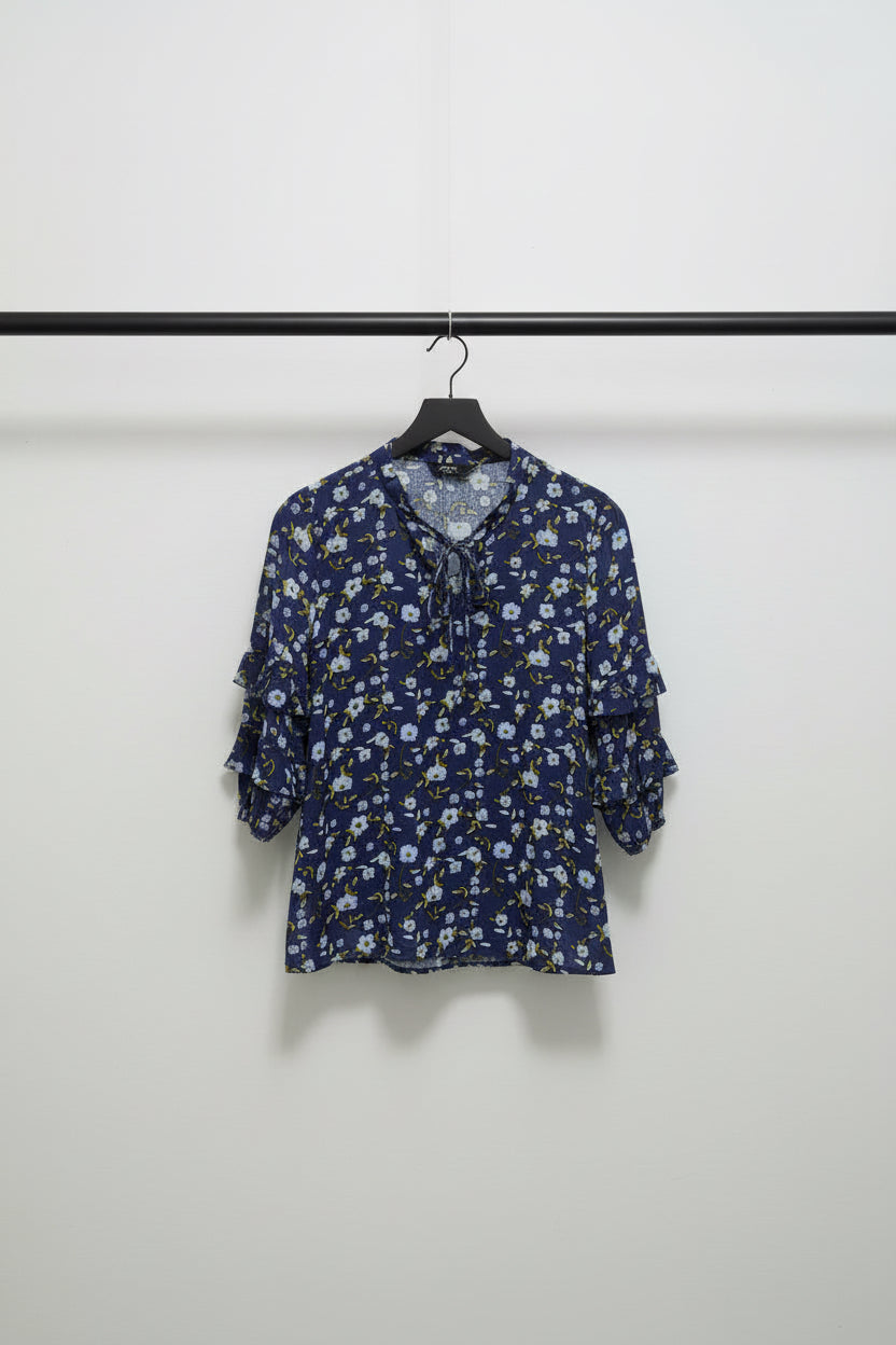 A women's long-sleeve blouse with a ruffled collar and a dense floral print, laid flat on a light gray fabric background. The base color is navy blue, with white and light blue flowers and small yellow accents.