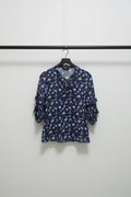 A women's long-sleeve blouse with a ruffled collar and a dense floral print, laid flat on a light gray fabric background. The base color is navy blue, with white and light blue flowers and small yellow accents.