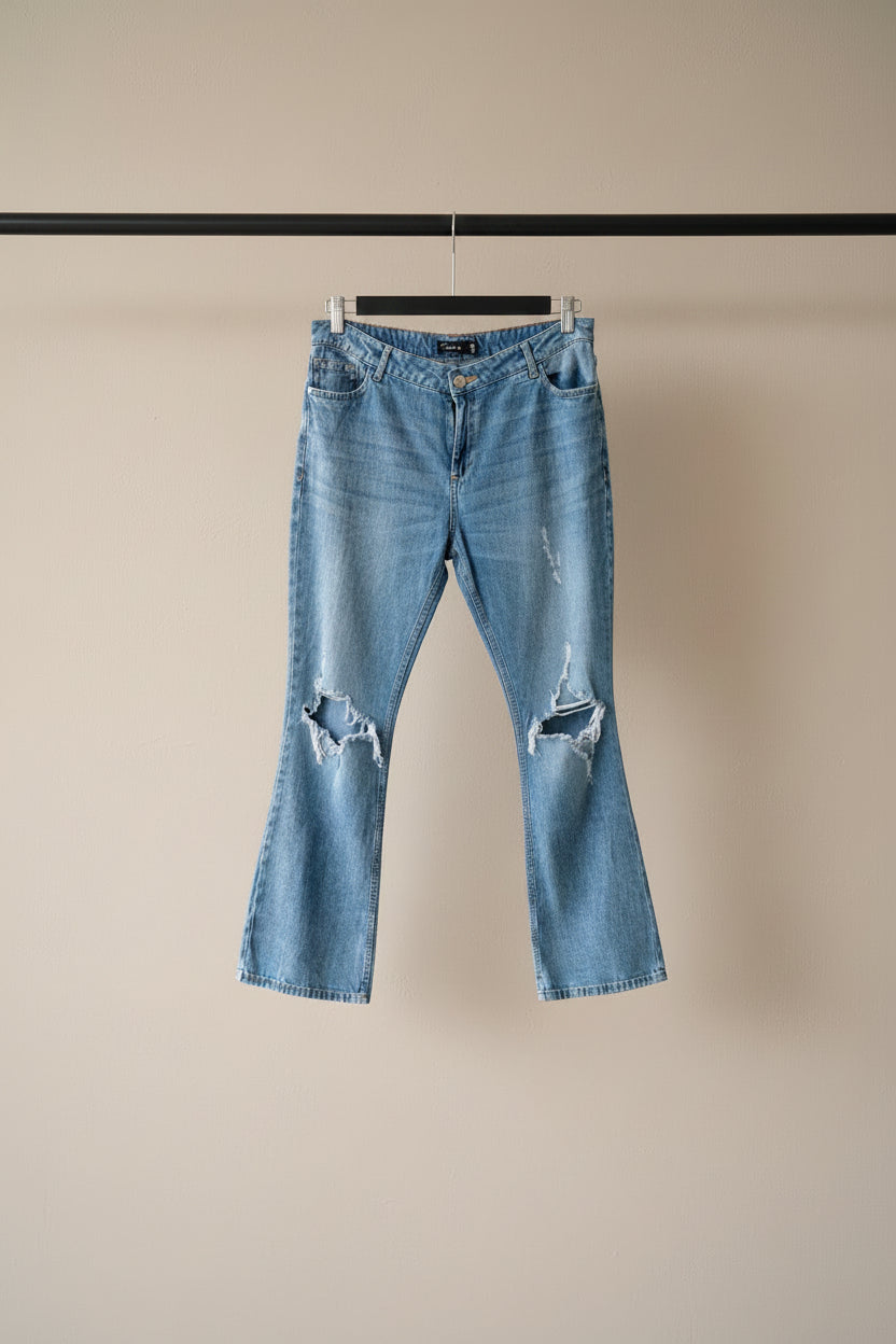 Light blue distressed denim jeans with ripped details on the thighs, hanging on a wooden hanger against a gray fabric backdrop.